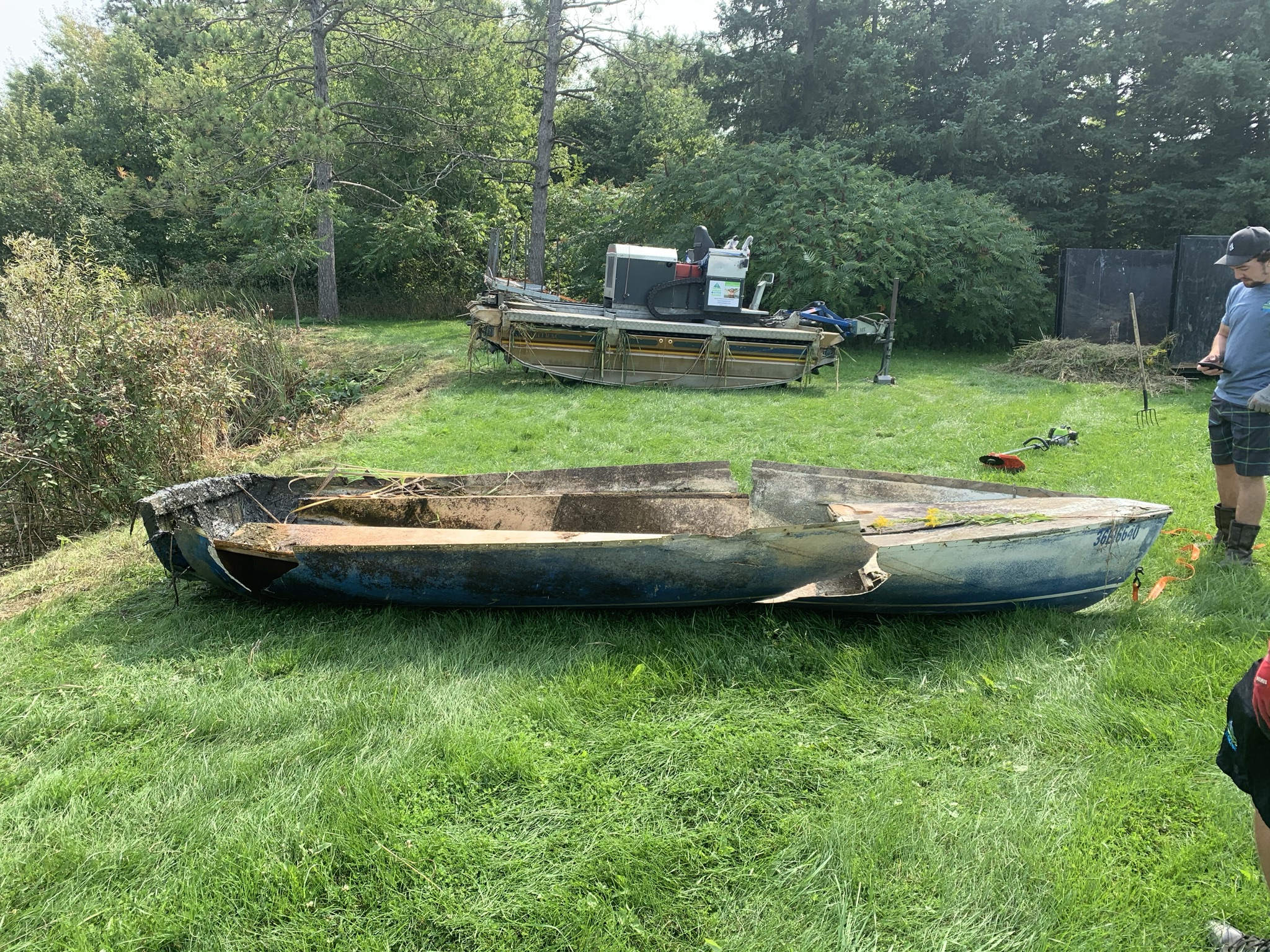 A person stands near a damaged, overturned boat on grassy land, surrounded by trees, tools, and equipment in a rural setting.