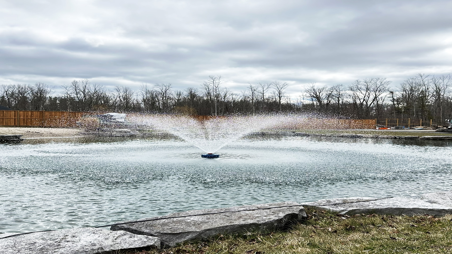 A fountain sprays water in a pond surrounded by trees and rocks on a cloudy day, with a wooden fence in the background.