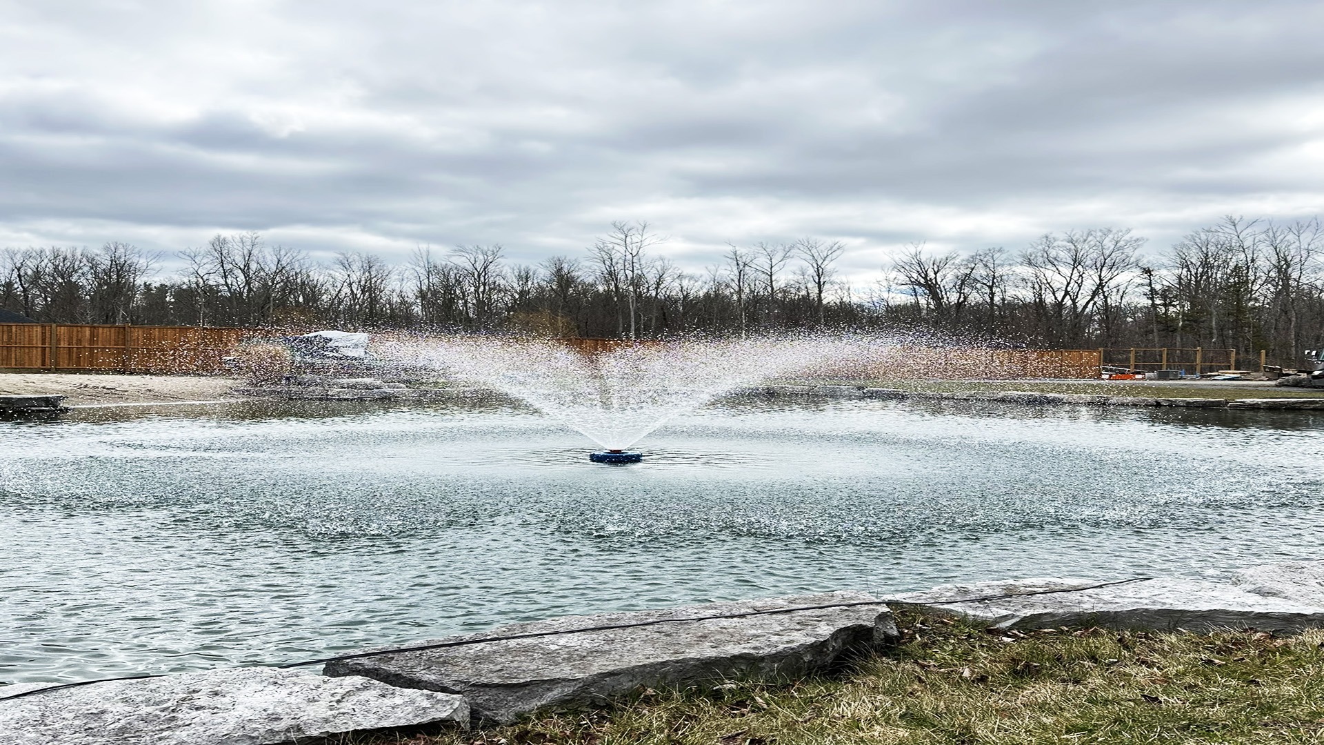 A pond with a central fountain spraying water, surrounded by bare trees, grass, and a wooden fence under a cloudy sky.
