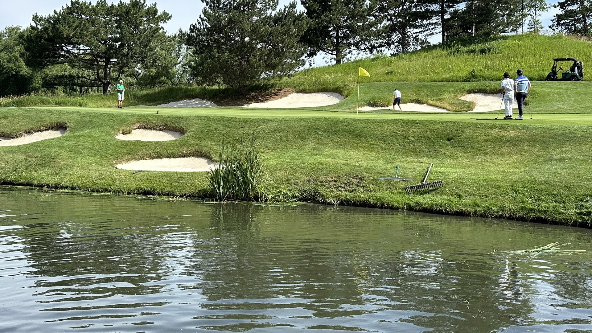 Four people playing golf near water, surrounded by sand traps and trees. A golf cart is visible on the right side.