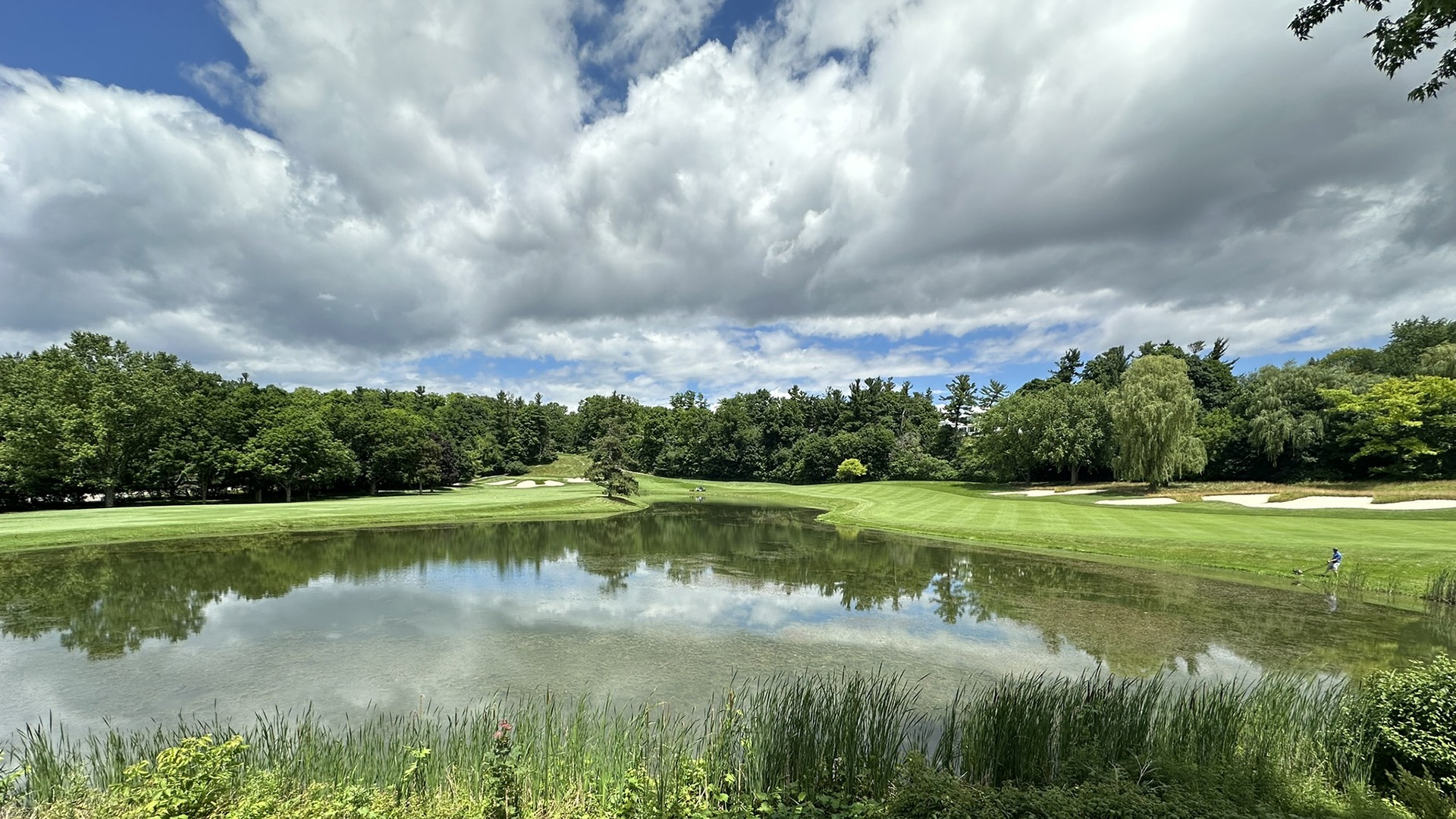 A serene golf course with lush greenery, reflecting pond, and cloudy sky. A person is walking near the water's edge.