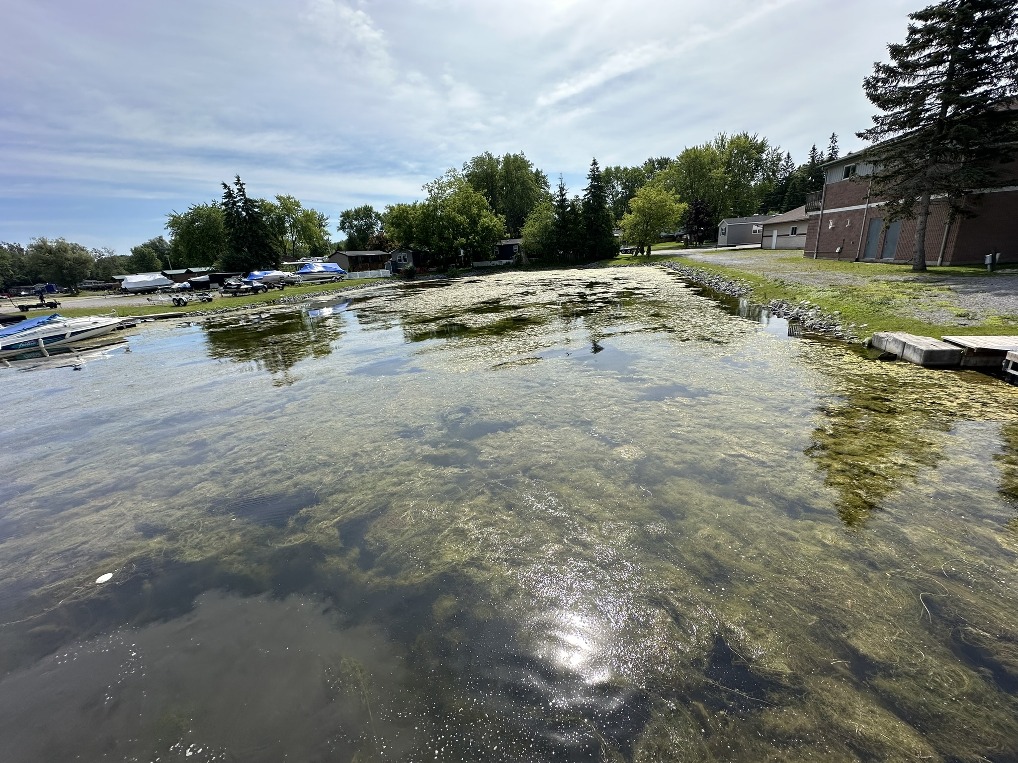 A serene lakeside scene with boats, clear water, surrounding trees, and a brick building. Overcast sky and calm atmosphere dominate the landscape.
