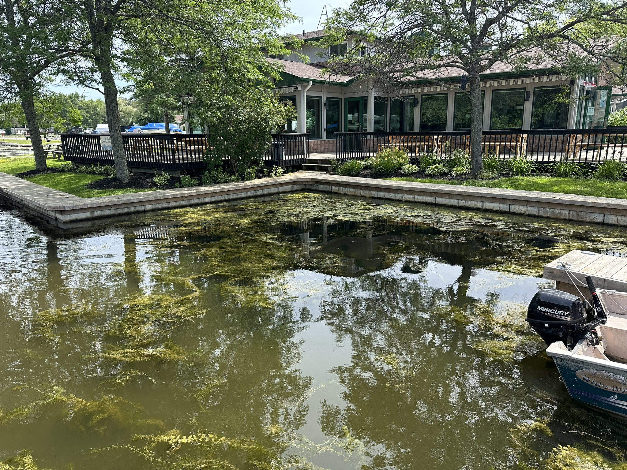 A canal lined with trees and vegetation, featuring a small dock and building with windows. A boat with an outboard motor is docked.