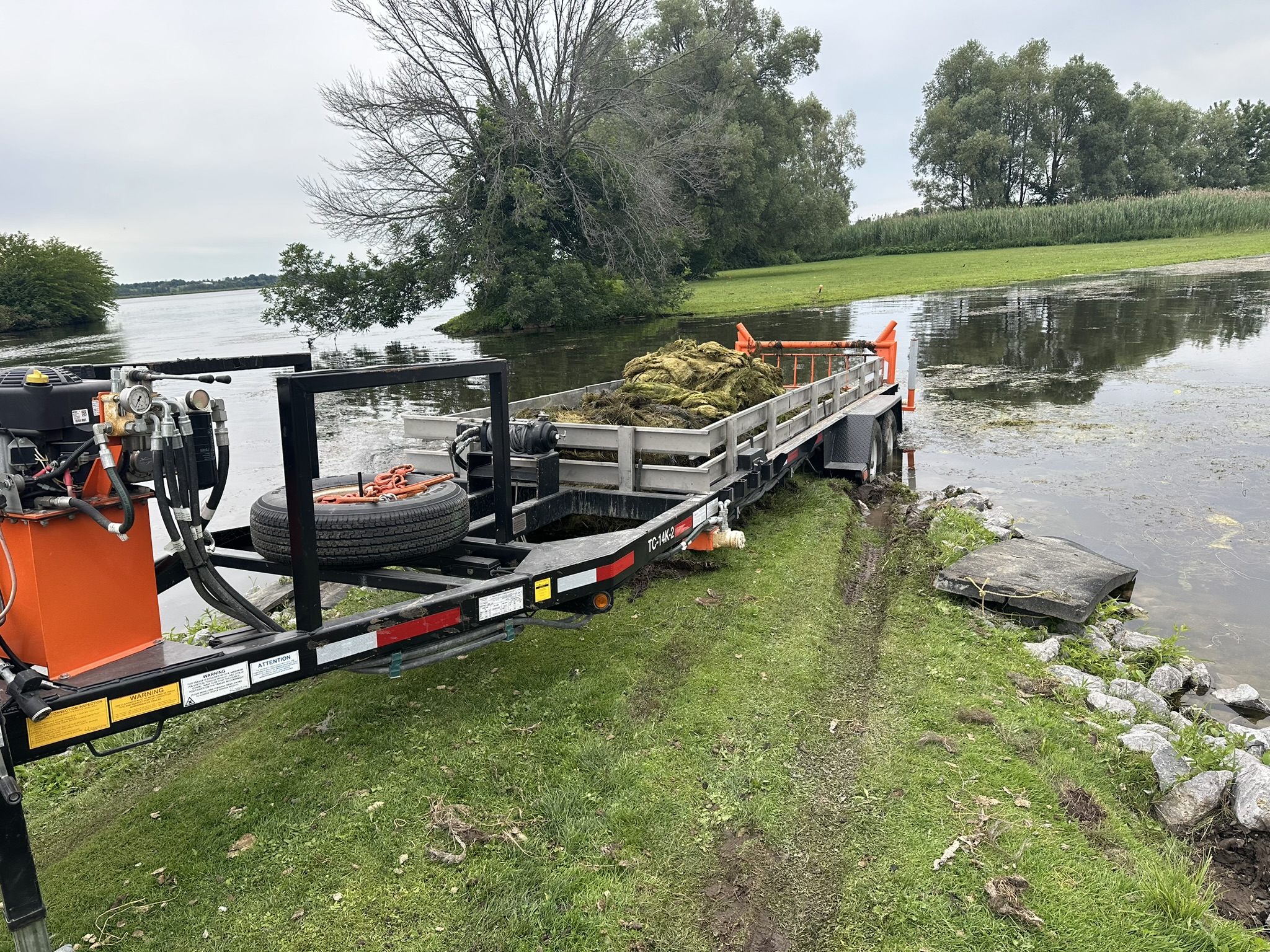 A trailer filled with aquatic vegetation sits by a tranquil lake, surrounded by lush trees and a grassy shoreline, under a cloudy sky.