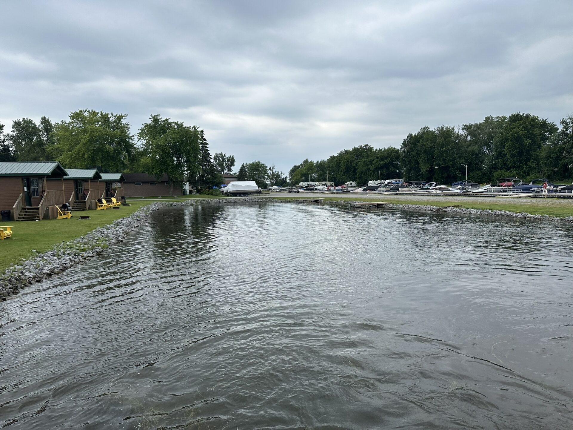 Lakeside scene with cabins, boats docked along the shore, and trees under a cloudy sky. No recognizable landmarks or historical buildings visible.