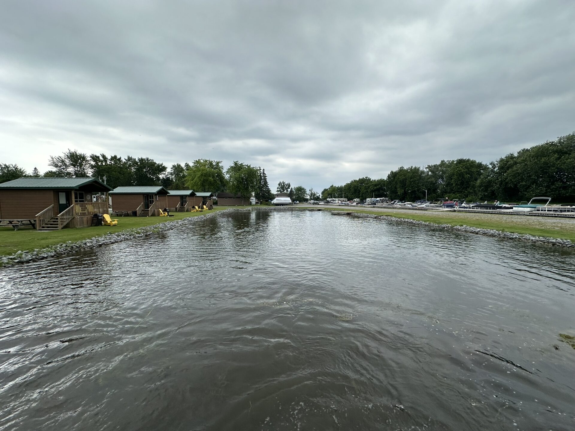 Row of cabins by a calm lake under a cloudy sky, flanked by trees and parked boats, creating a peaceful atmosphere.