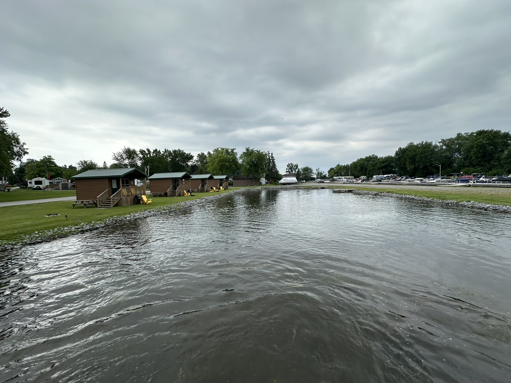 Lakeside view with several cabins, parked cars, and boats. Cloudy sky and calm water create a serene atmosphere.