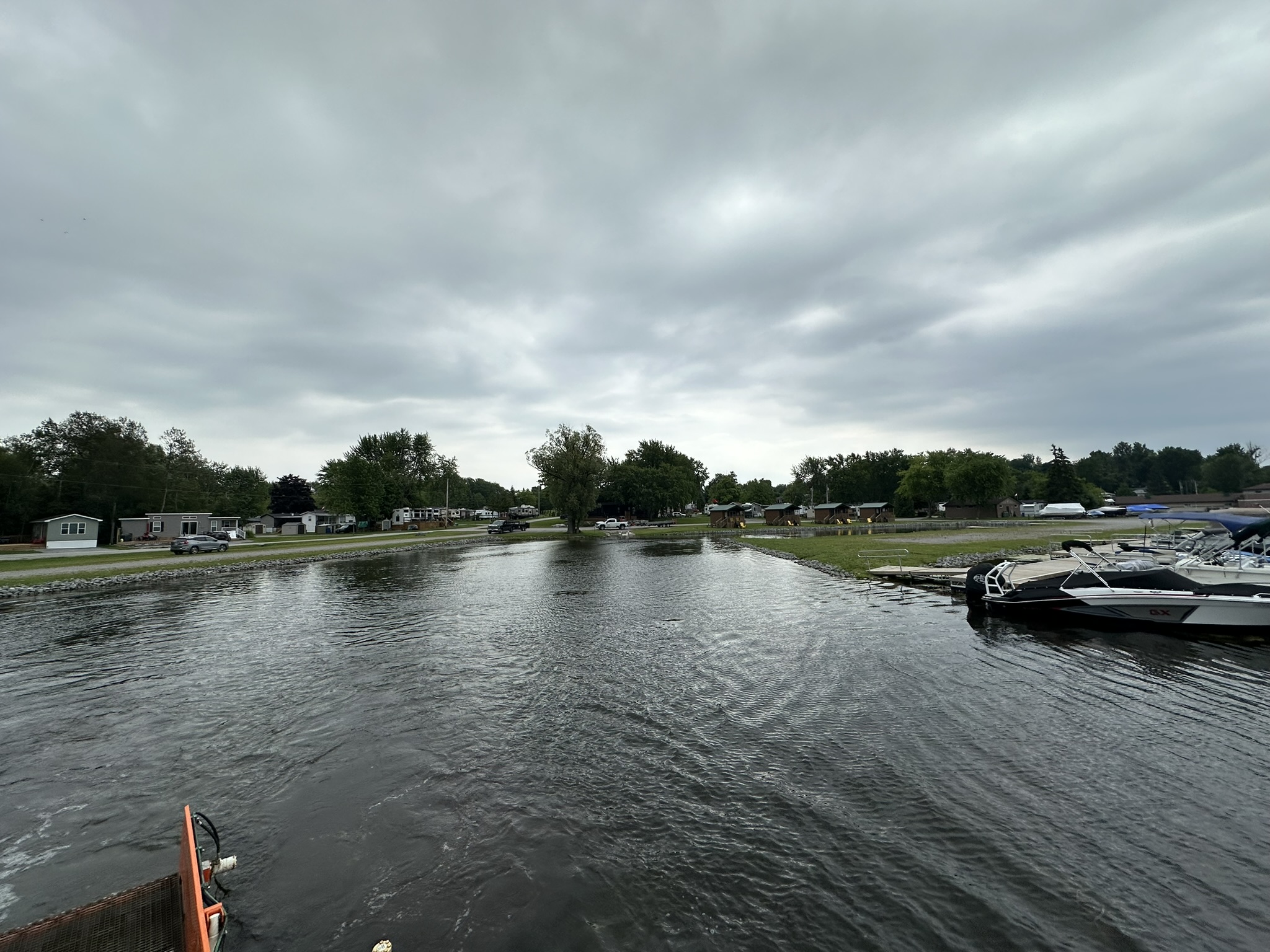 A tranquil lakeside scene with boats docked, surrounded by trees and houses, under a cloudy sky, creating a serene atmosphere.