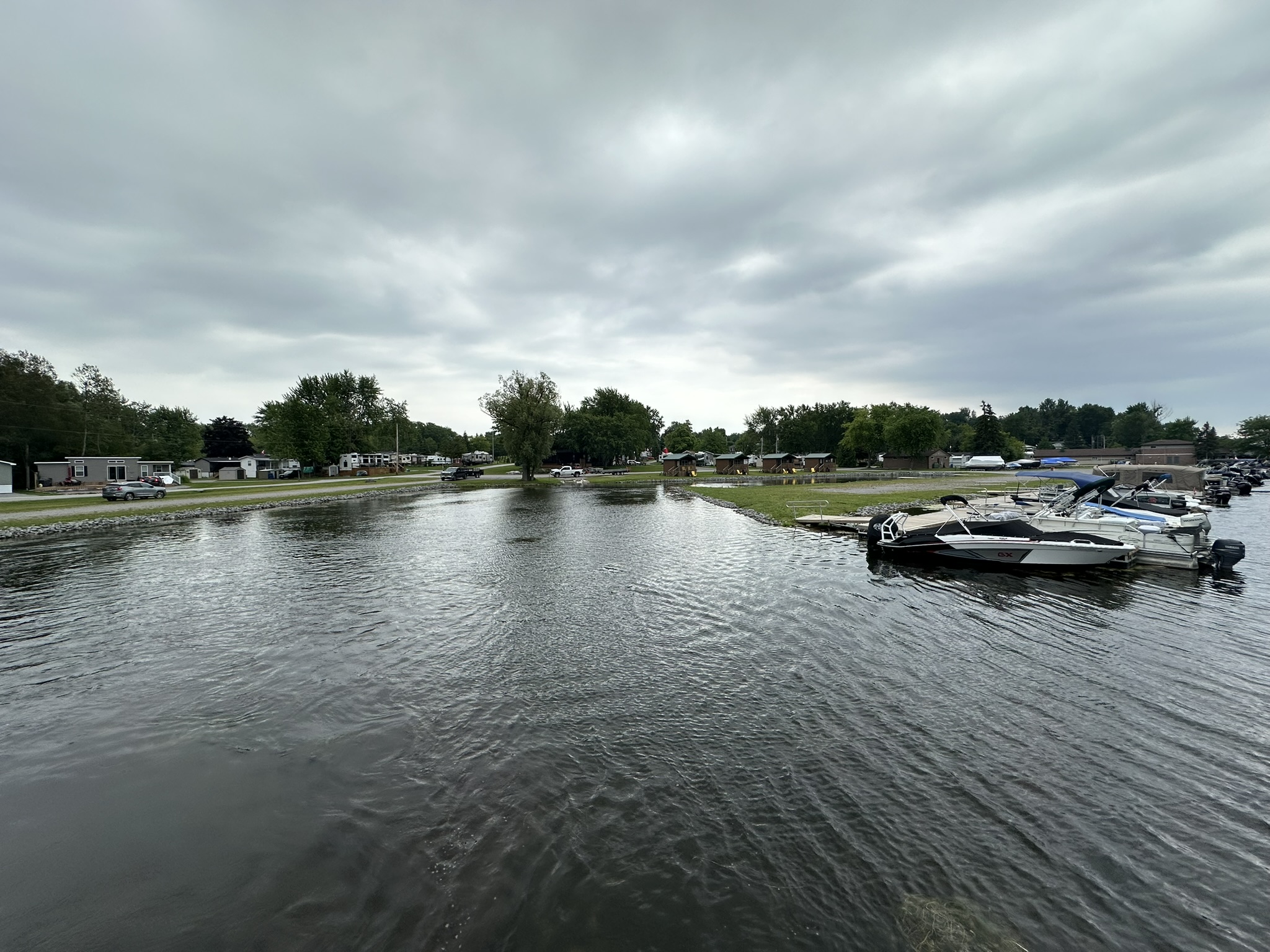 A cloudy sky overlooks a marina with parked boats on calm water, surrounded by trees and residential buildings along the shore.