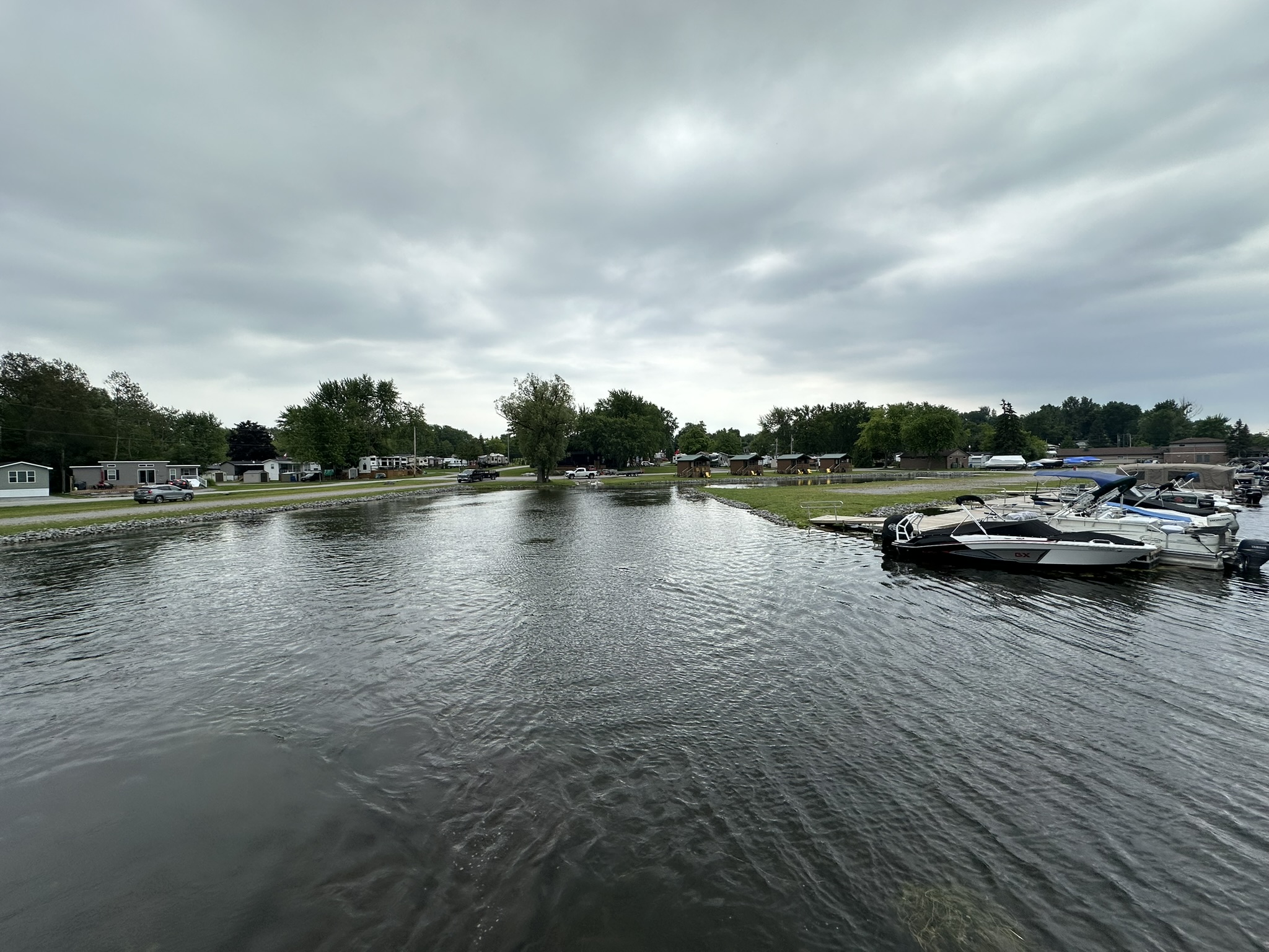 Cloudy sky over a calm marina with several boats docked, surrounded by trees and buildings, creating a peaceful and serene environment.