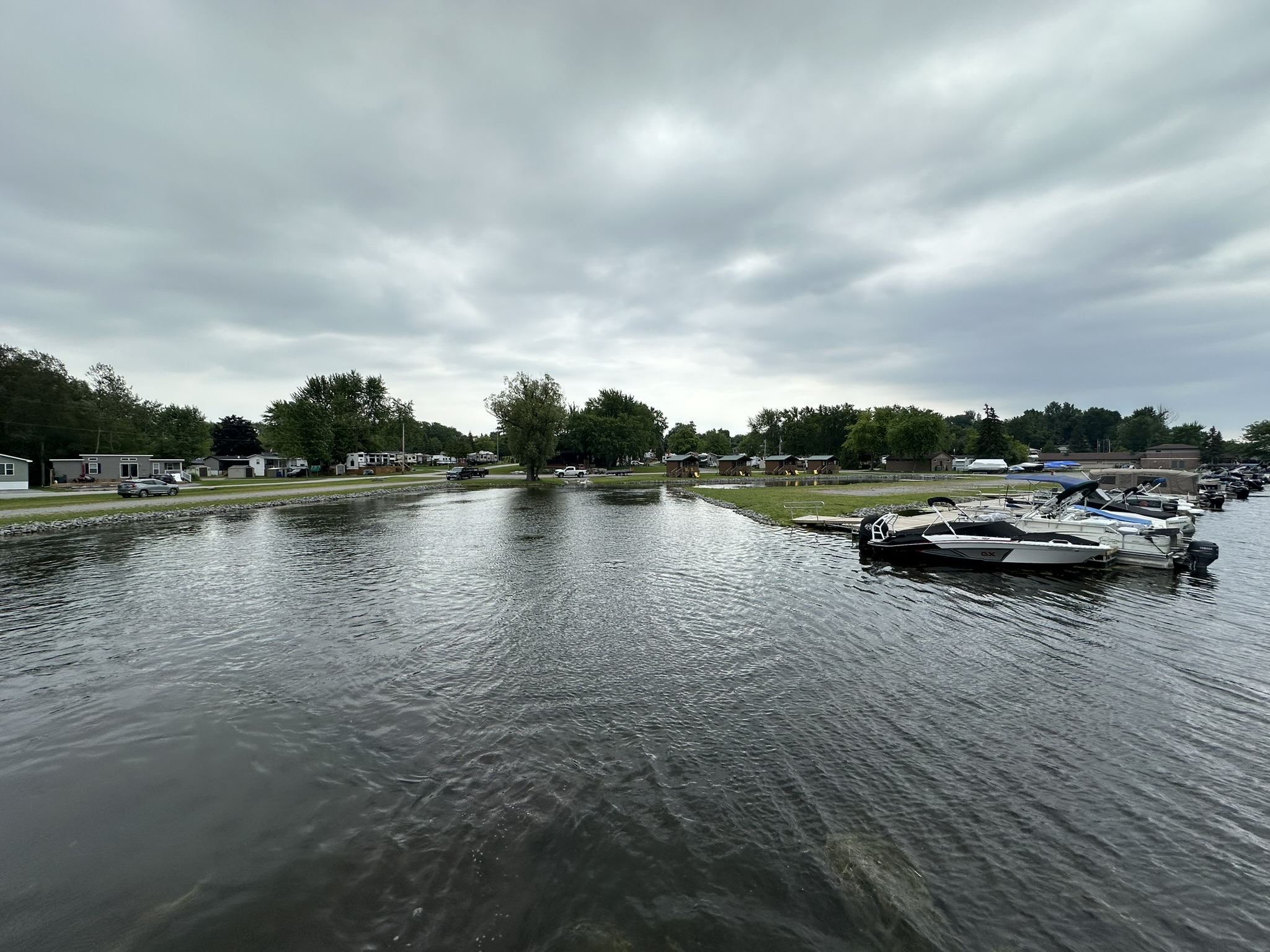 Boats docked by a calm riverbank, with houses and trees under a cloudy sky, creating a peaceful, rural scene.