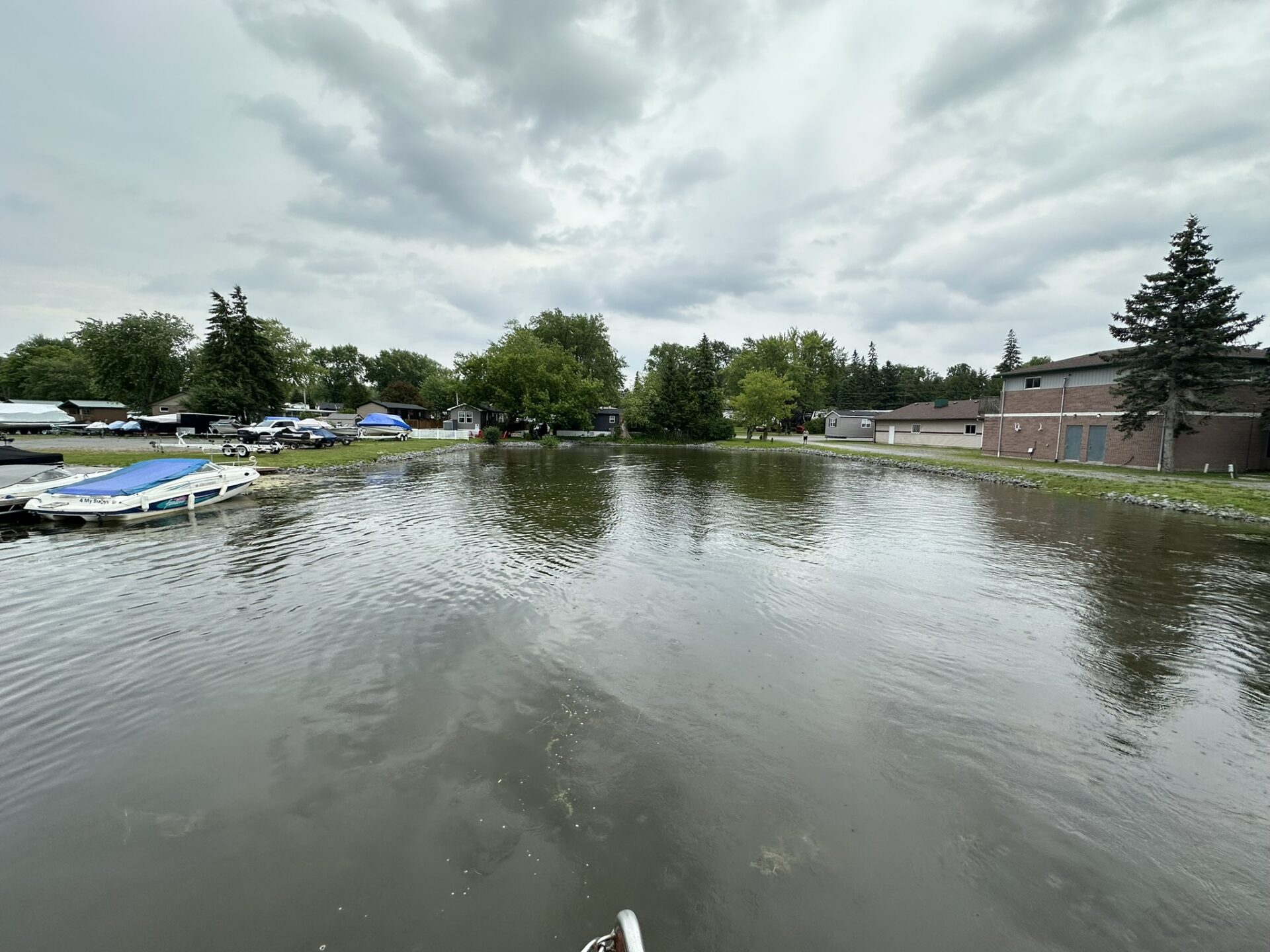 A calm waterfront scene features parked boats, a building, trees, and a person walking along the shore under a cloudy sky.