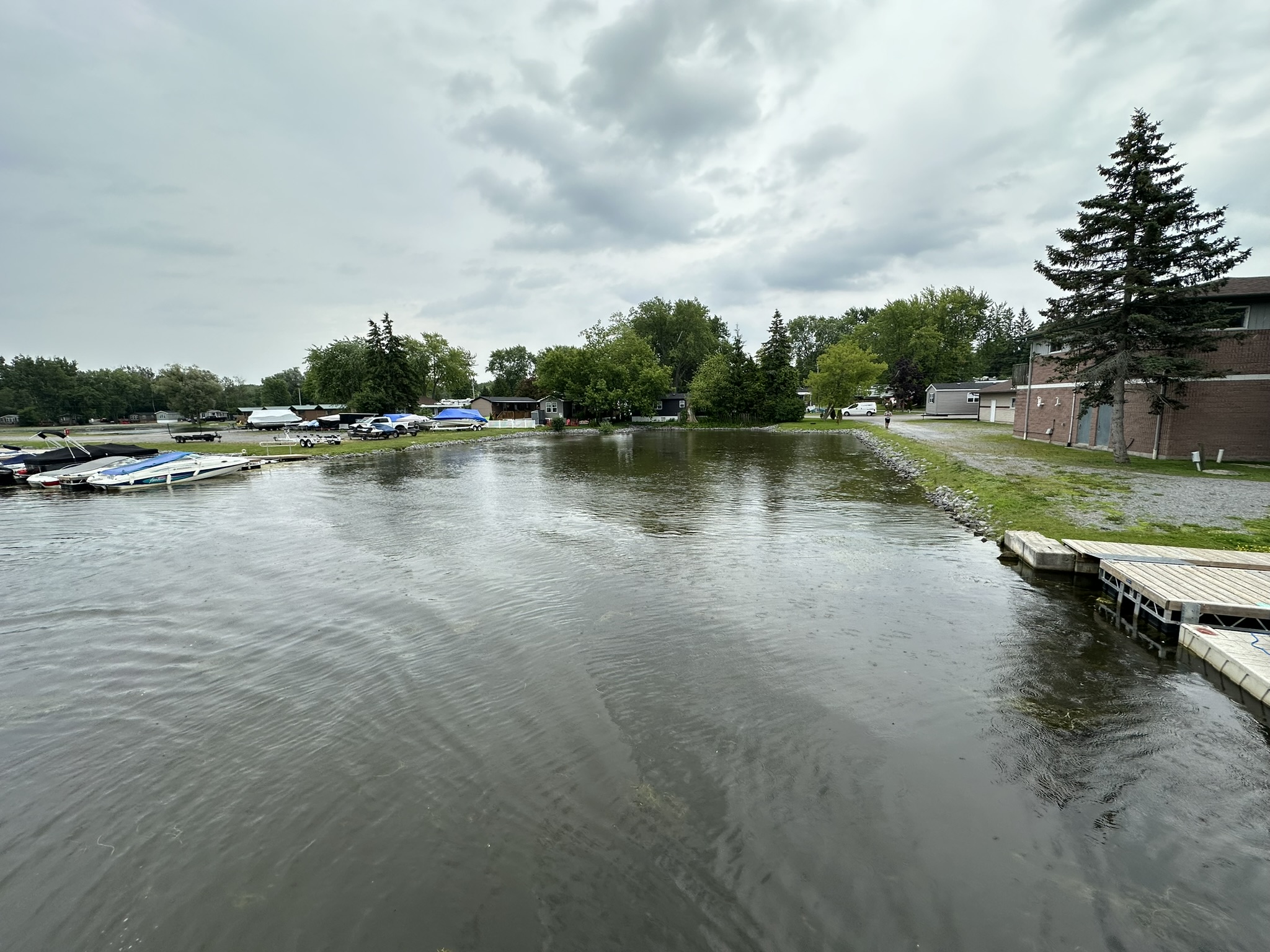 A cloudy lakeside view featuring docks, boats, and buildings along the shore, surrounded by trees and a gravel pathway.