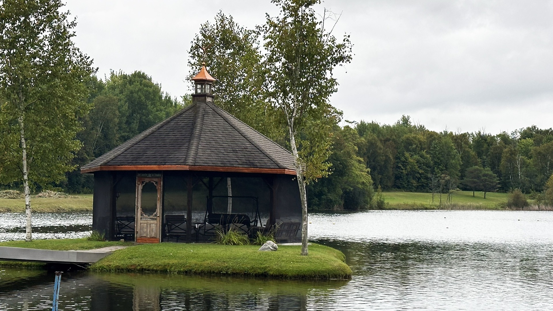 A small gazebo sits on an island in a serene lake, surrounded by lush trees under a cloudy sky.