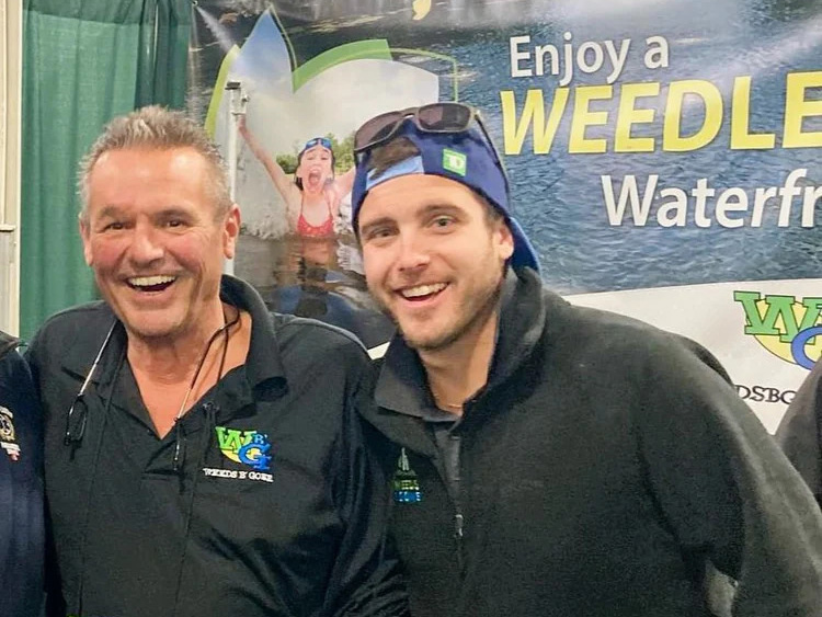 Two people smiling in front of a booth with a "Enjoy a WEEDLE Waterfront" banner, wearing casual attire and sunglasses.