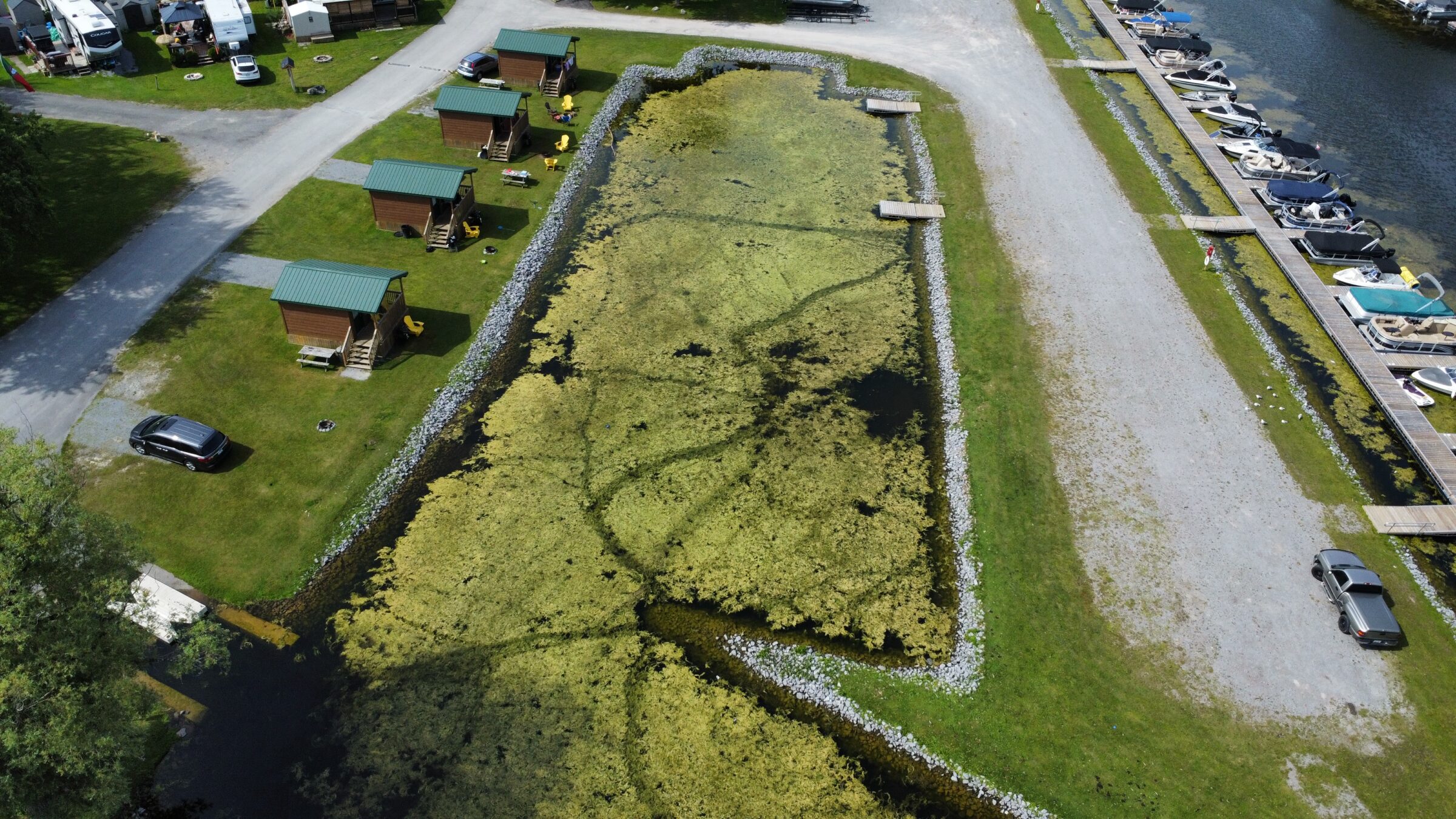 Aerial view of a campground with cabins, parked cars, and a marina with boats beside a rectangular pond overgrown with algae.
