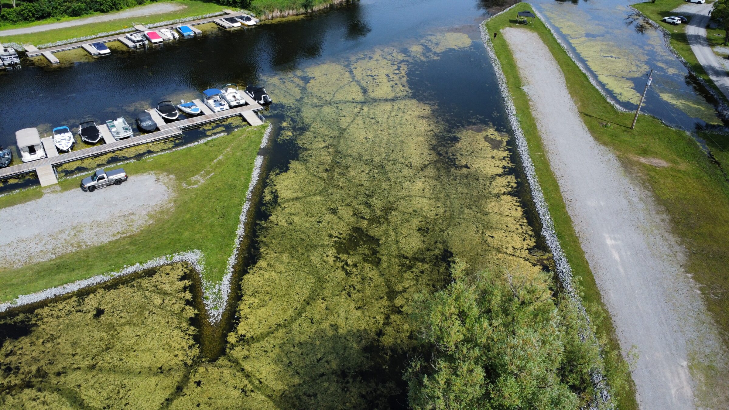 Aerial view of a marina with parked boats, grassy paths, gravel roads, and a parked truck near the waterway.