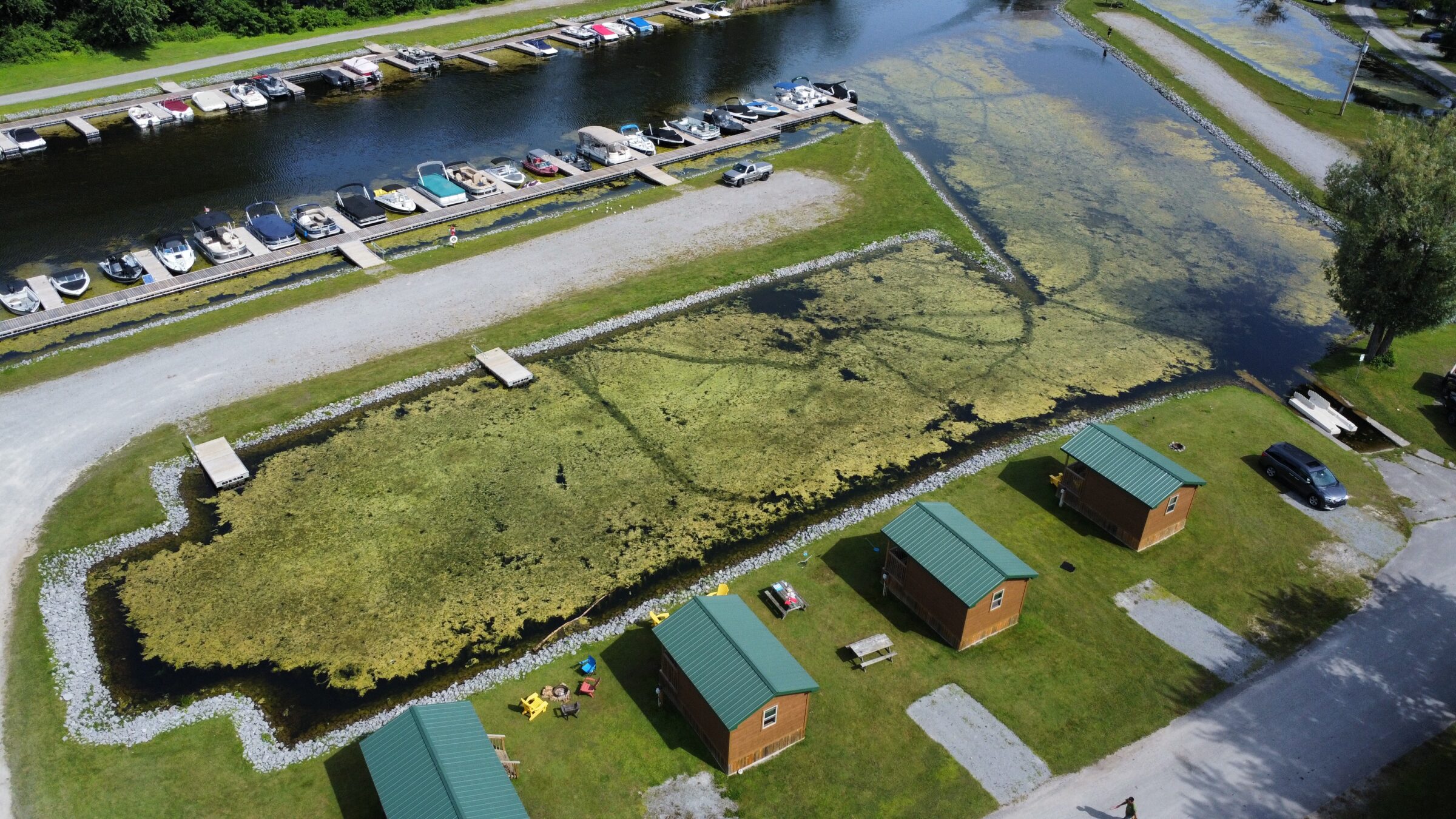 Aerial view of a marina with docked boats, nearby grassy area with cabins, and a person walking on the path alongside.