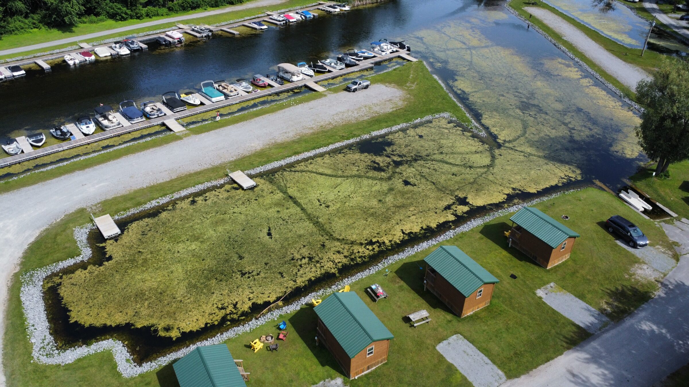 Aerial view shows small cabins with green roofs, parked cars, and a row of docked boats along a canal with grassy areas.