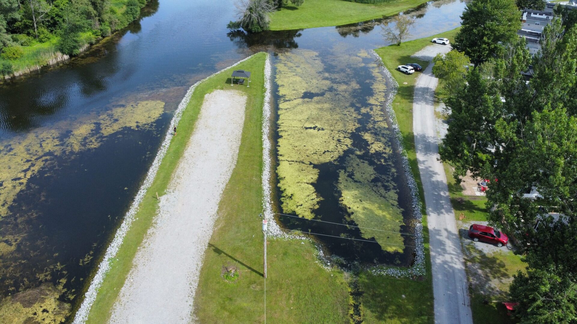 Aerial view of a narrow waterway bordered by grass and trees, with cars parked nearby. A lone person strolls along the path.