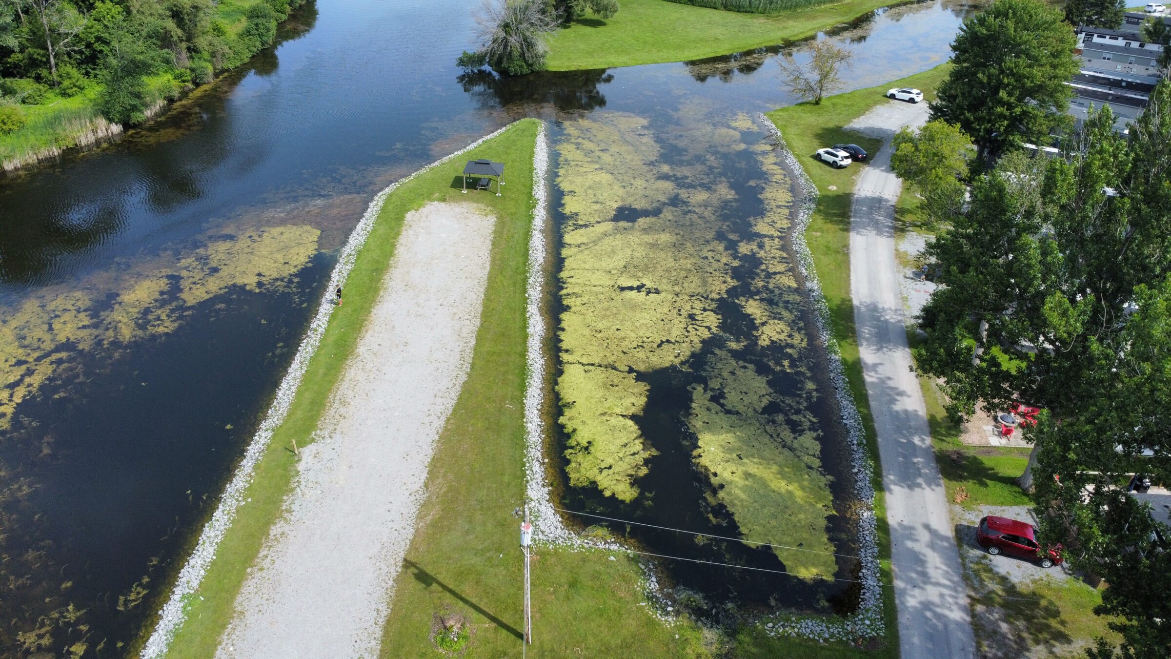 Aerial view of a narrow peninsula with greenery, a road, parked cars, and water on both sides under a clear sky.