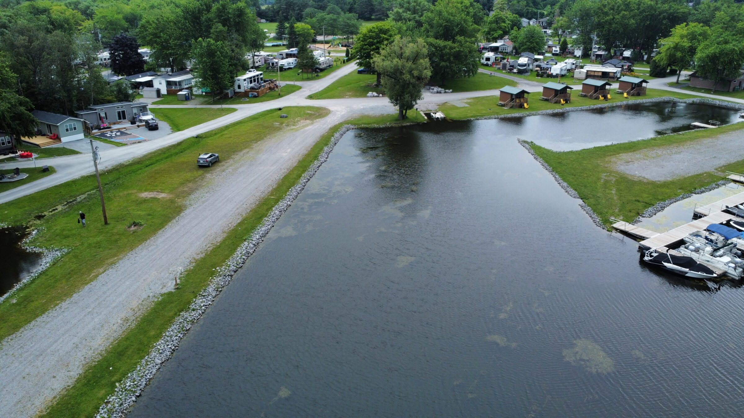Aerial view of a lakeside residential area with small cabins, parked vehicles, a person walking, and boats docked at a marina.