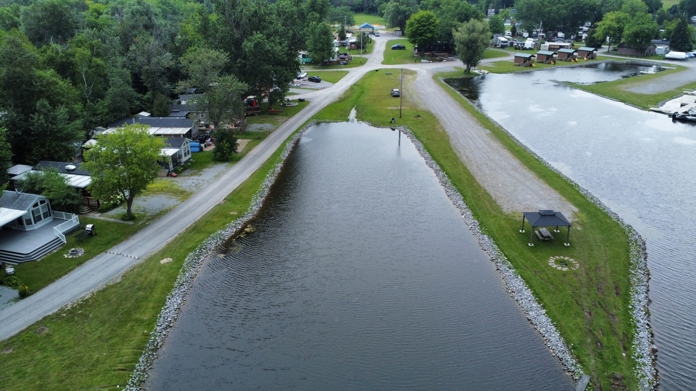 Aerial view of a serene lakeside community with cabins, roadways, trees, and small docks. Picnic area with a shelter by the water.