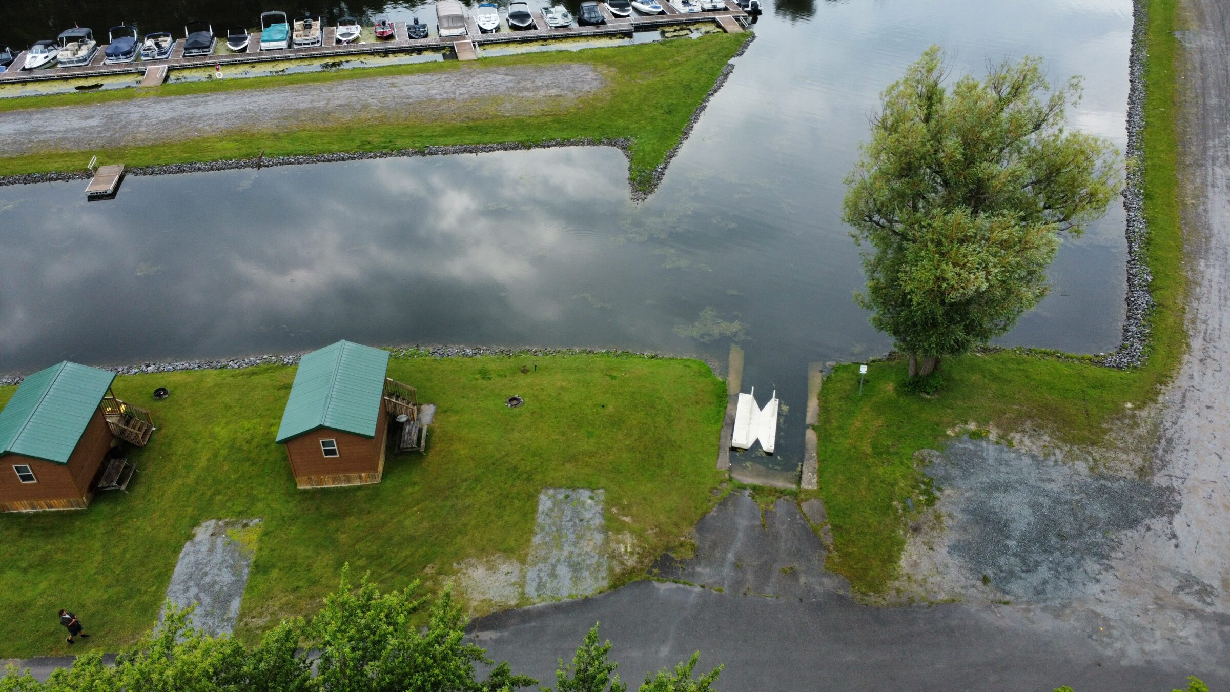 Aerial view of a marina with docked boats, green-roofed cabins, a pond, and a surrounding grassy area. A person walks along the path.