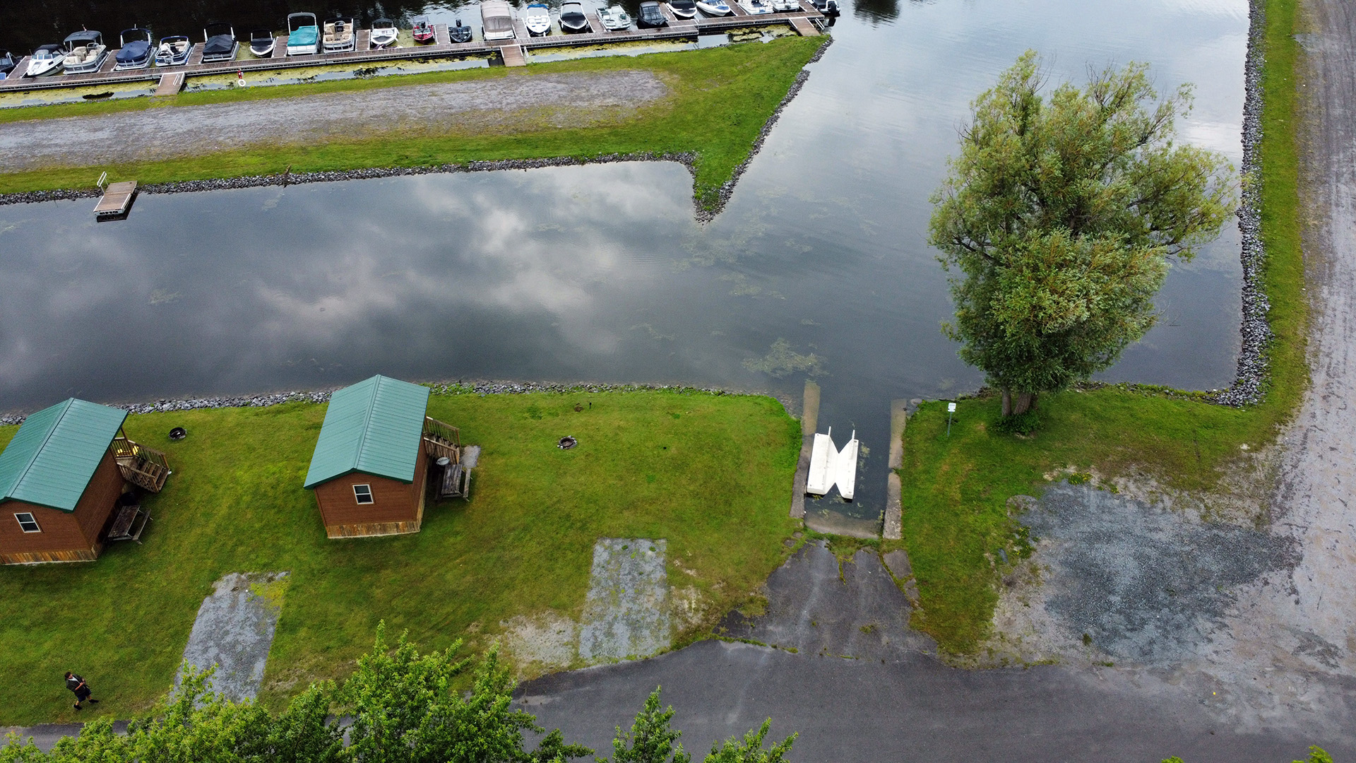 Aerial view of a marina with parked boats, two small cabins, grassy areas, a tree, and a person walking along a path.