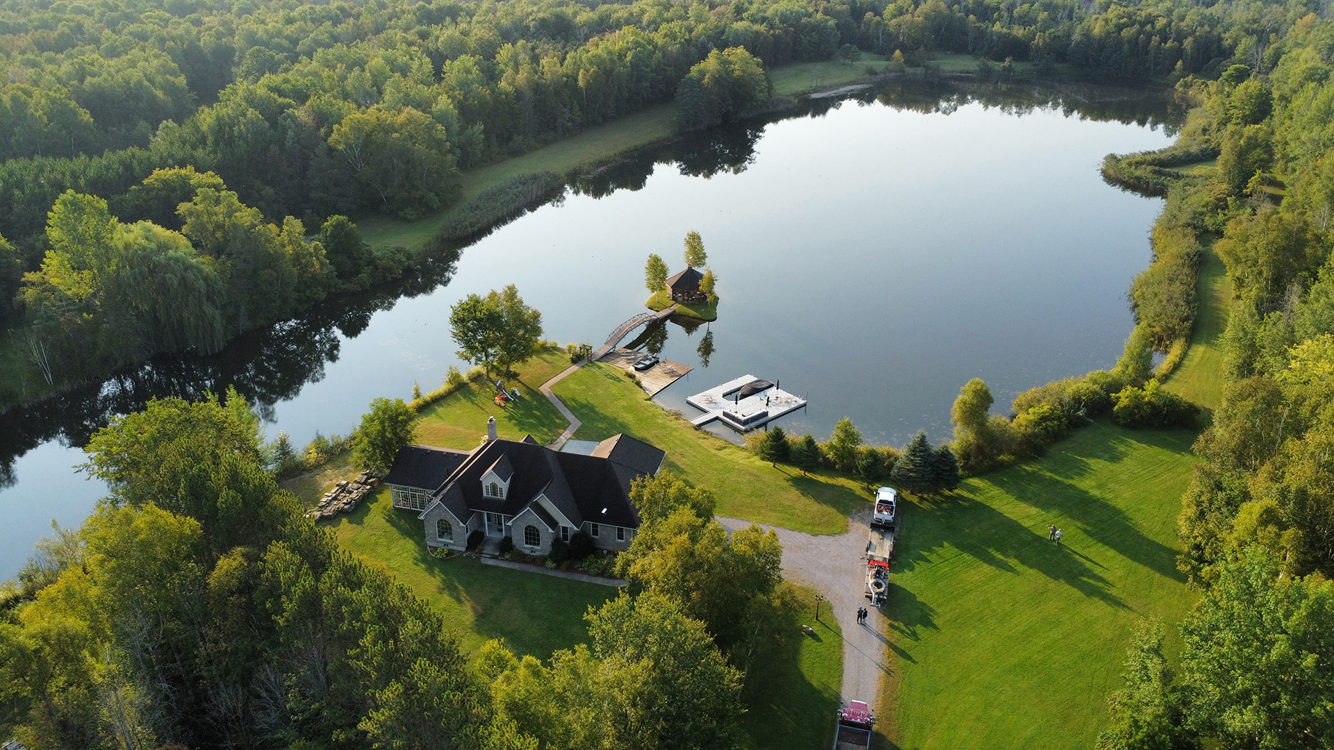 A serene lakeside scene with a house, dock, and cars surrounded by lush greenery, reflecting on the calm water.