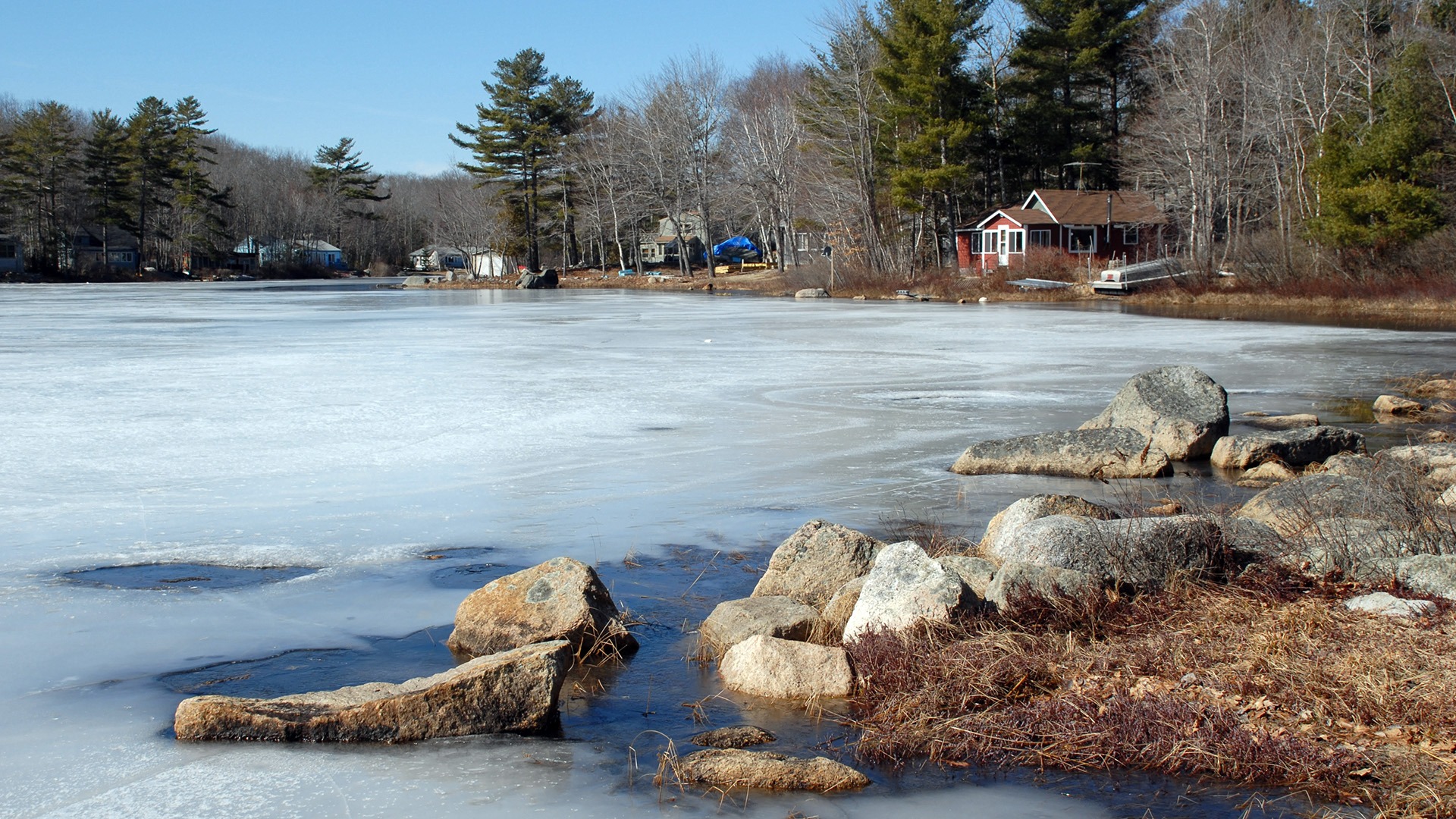 A serene, frozen lake bordered by rocks and evergreen trees, with small rustic cabins nestled among the forested shoreline under a clear blue sky.