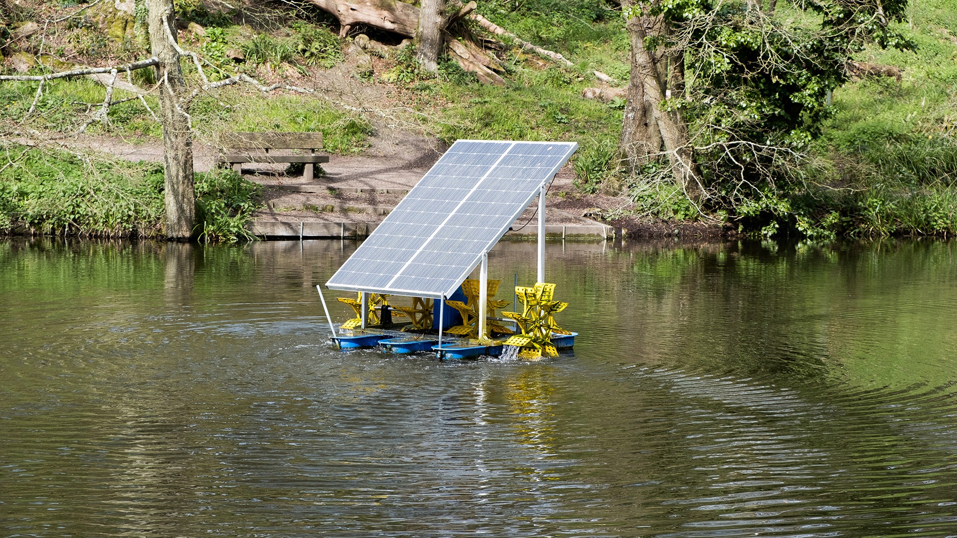 A floating solar-powered device with yellow paddles moves on a calm pond, surrounded by lush green trees and a wooden bench nearby.