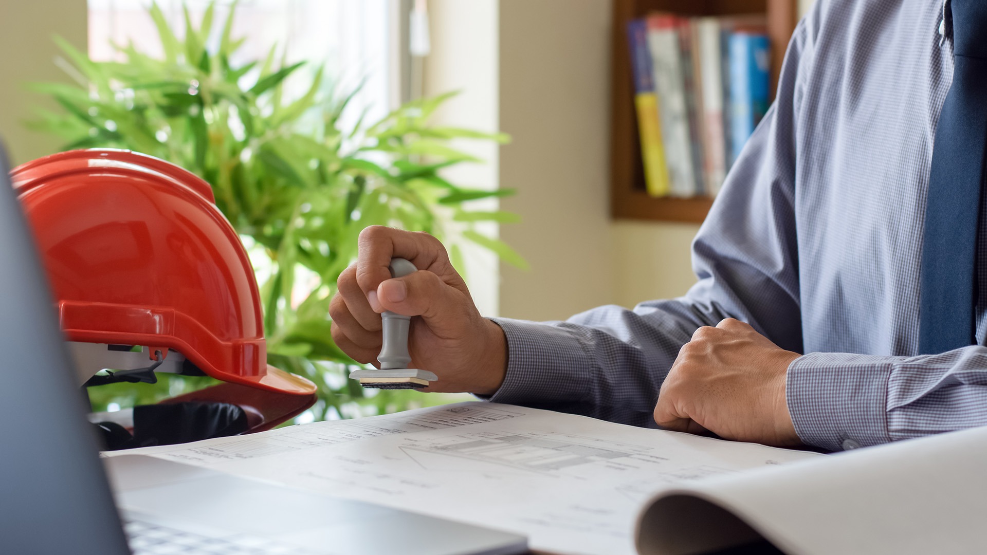A person stamps documents in an office setting with a desk, red hard hat, and plant visible.