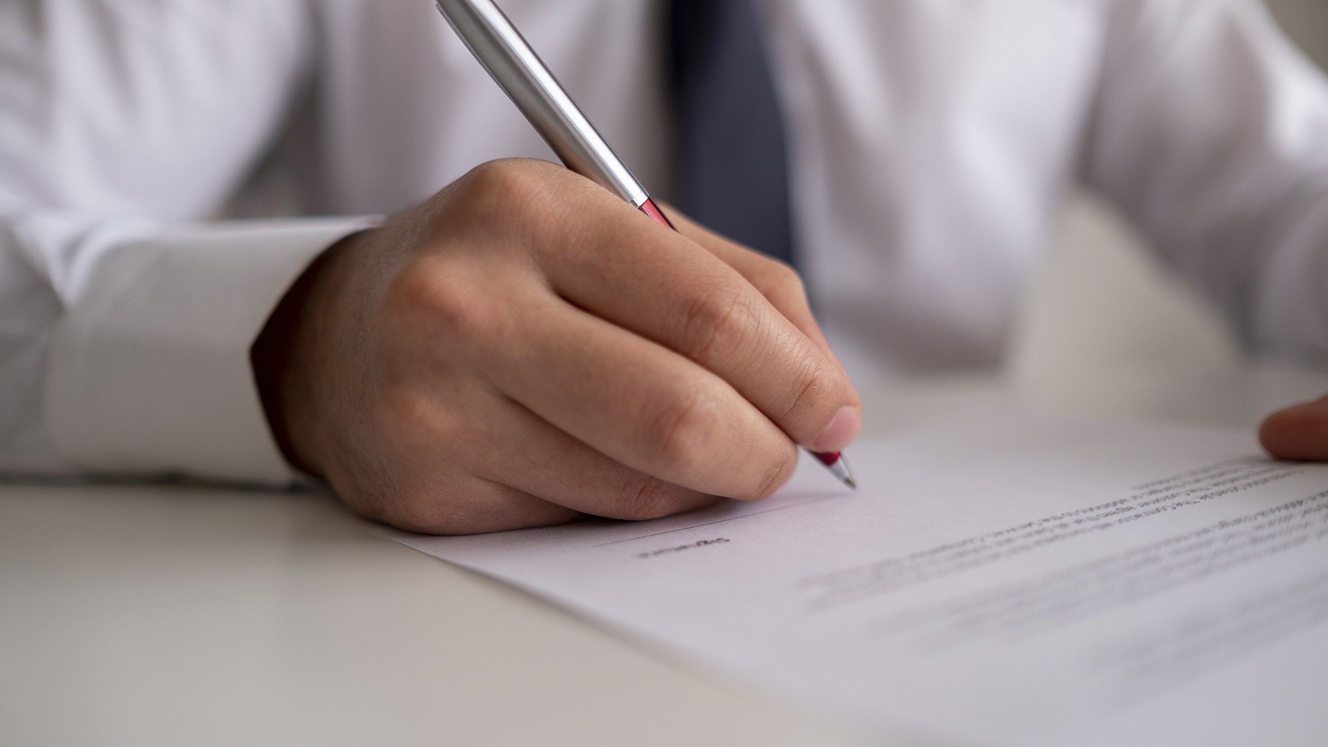 A person, dressed formally, is writing on a piece of paper with a pen, focusing on precise handwriting in a professional setting.