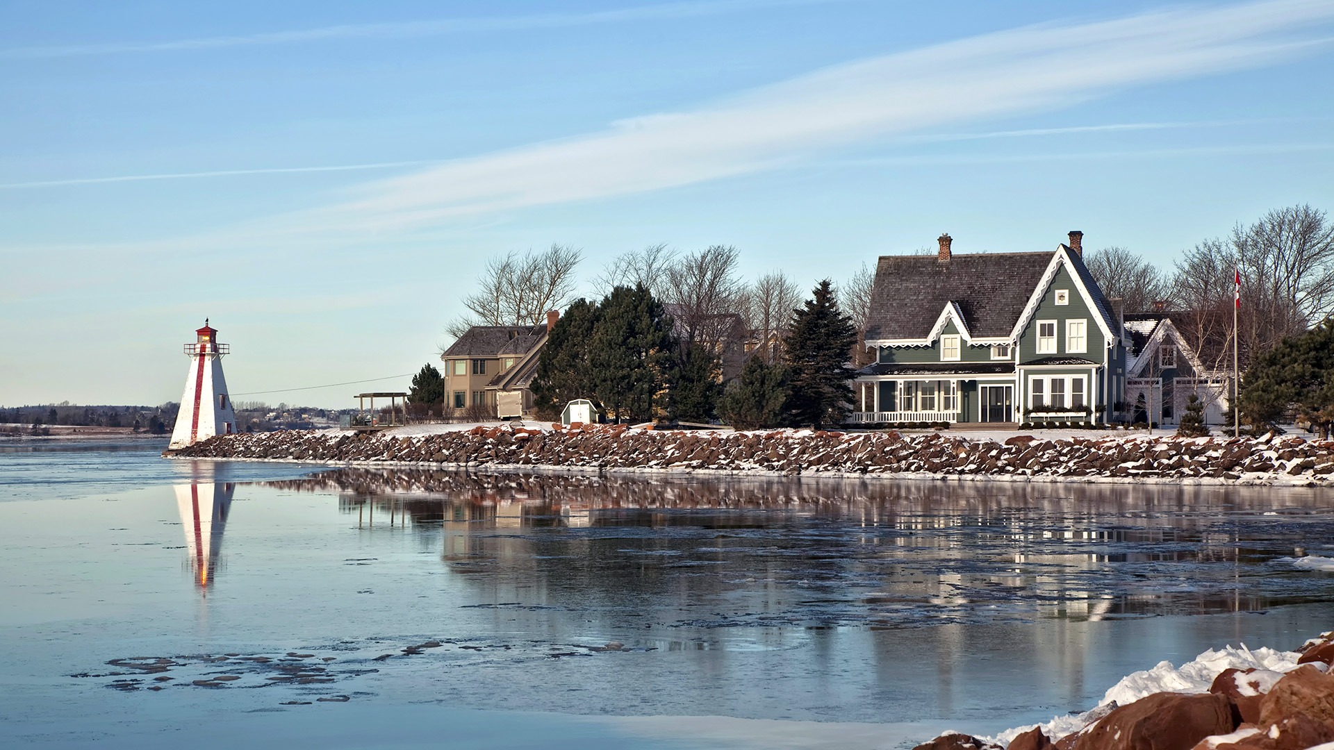 Snowy shoreline features a lighthouse and charming houses. Reflective icy water complements the serene winter landscape, accented by sparse trees and calm skies.