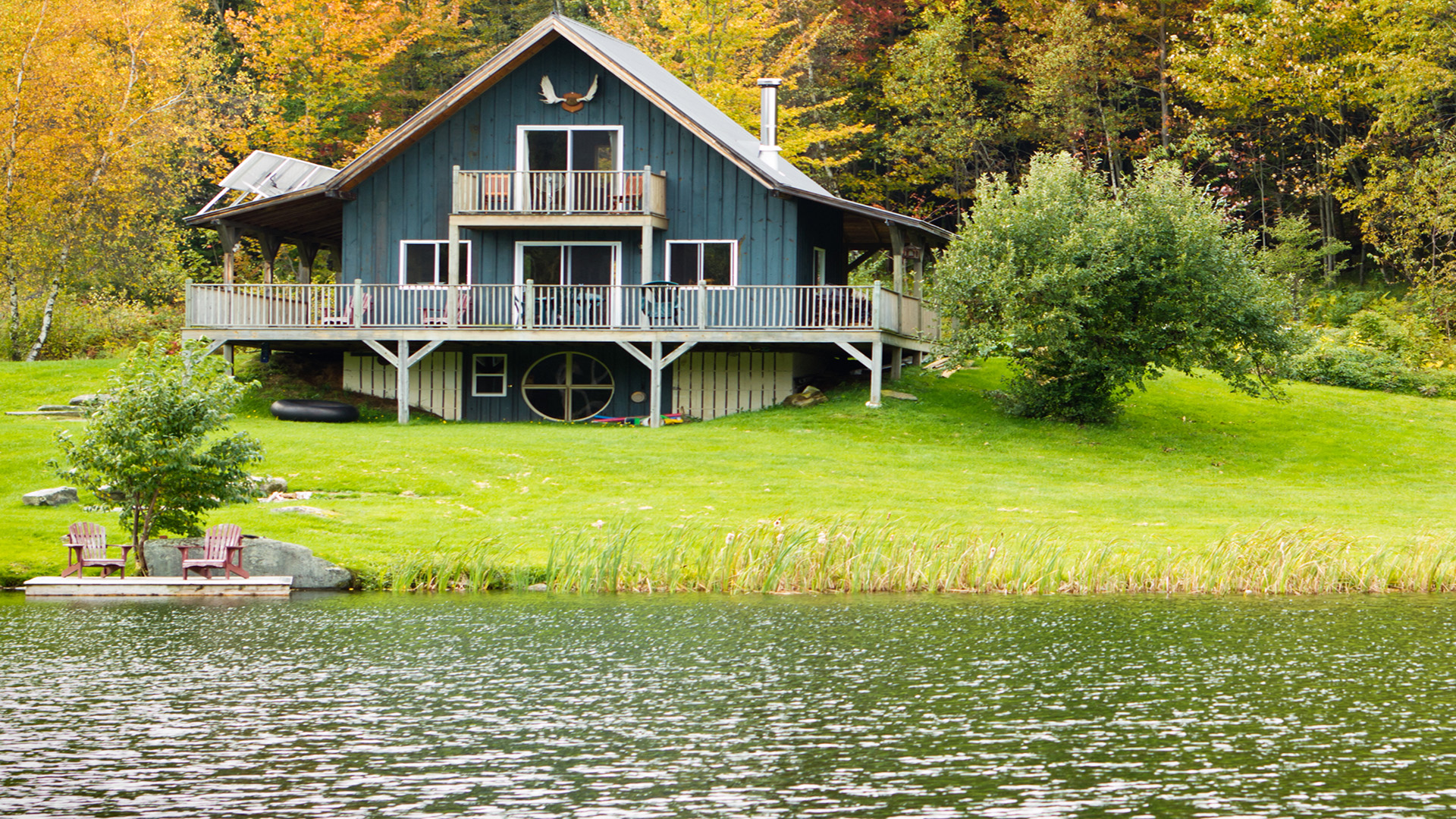 A cabin by the lake surrounded by autumn trees, with chairs on the shore and a tranquil, reflecting water surface.