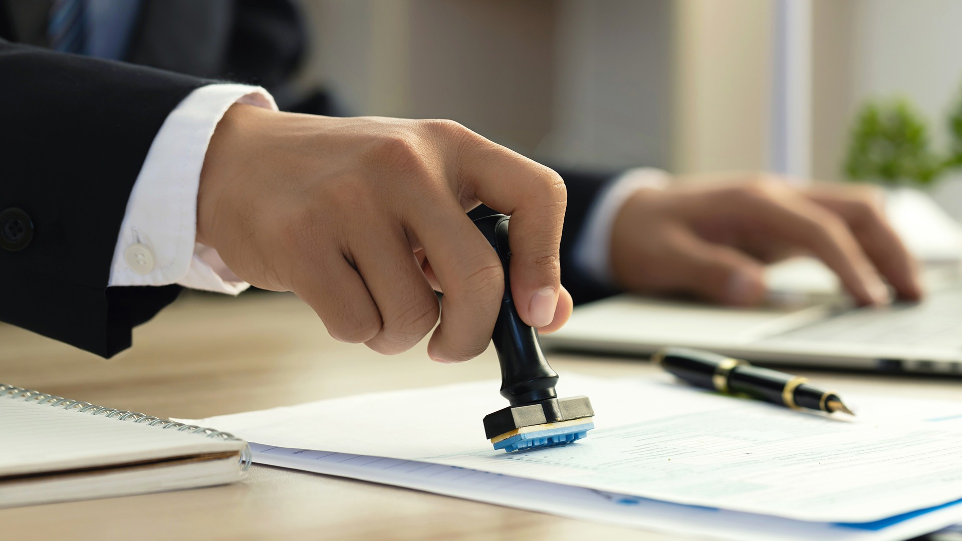 A person in formal attire stamps documents on a desk beside a pen, notepad, and laptop, suggesting an office environment.