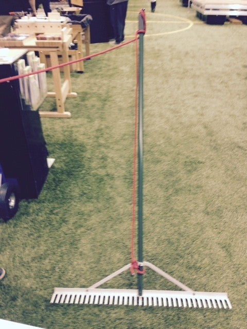 A rake is balanced on a green indoor field, with tables and equipment visible in the background. People are standing nearby.