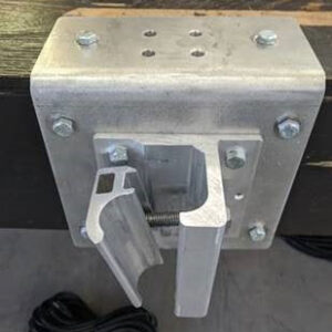 A metallic industrial bracket mounted on a wooden surface, featuring bolts and a hinged mechanism. Electrical cables are visible beneath the setup.
