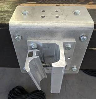 A metallic industrial bracket mounted on a wooden surface, featuring bolts and a hinged mechanism. Electrical cables are visible beneath the setup.