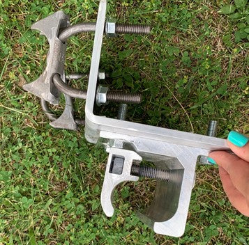 A person with teal nails holds a complex metal tool or clamp on grass, featuring various screws and adjustable components.