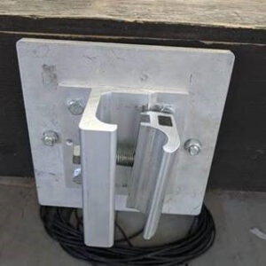 Metallic wall bracket mounted on a dark wooden surface, with coiled black cable beneath. Appears industrial, likely for securing or hanging items.