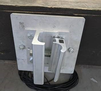 Metallic wall bracket mounted on a dark wooden surface, with coiled black cable beneath. Appears industrial, likely for securing or hanging items.