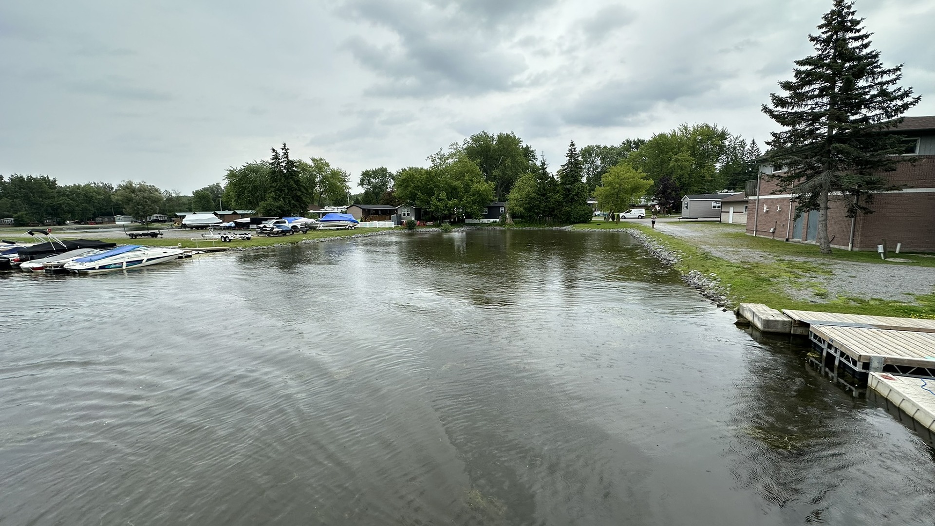 A calm waterfront scene with boats, trees, and buildings. A person walks near a brick building under a cloudy sky.