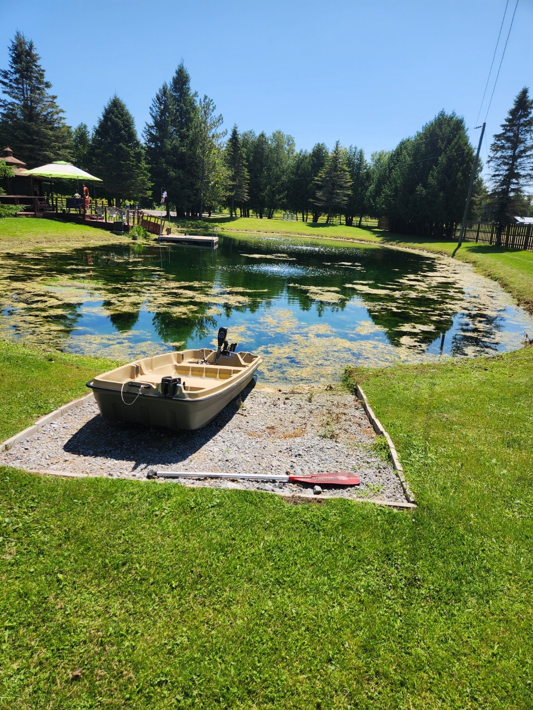 A small boat rests by a tranquil pond with a person nearby, surrounded by lush trees under a clear blue sky.